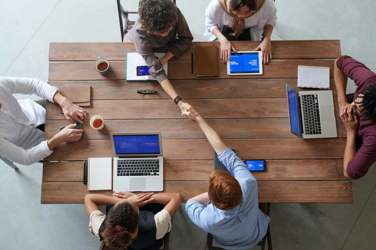 Business Team Seated Around a Table with Various Mobile Devices and Two People Reaching Out to Shake Hands