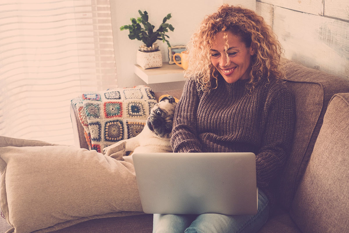 Smiling woman remote working from home with laptop computer on sofa with dog.