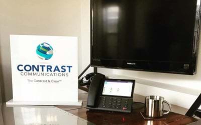 Contrast Communications Logo Sign on Desk with VoIP Phone and Wall Mounted Monitor in Background