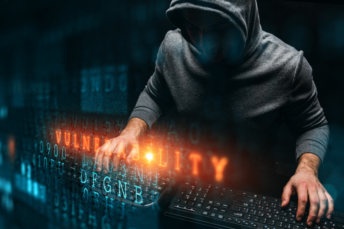 A hacker in hoodie using two keyboards at once with reddish text that says 'VULNERABILITY' overlaid on top on the image.