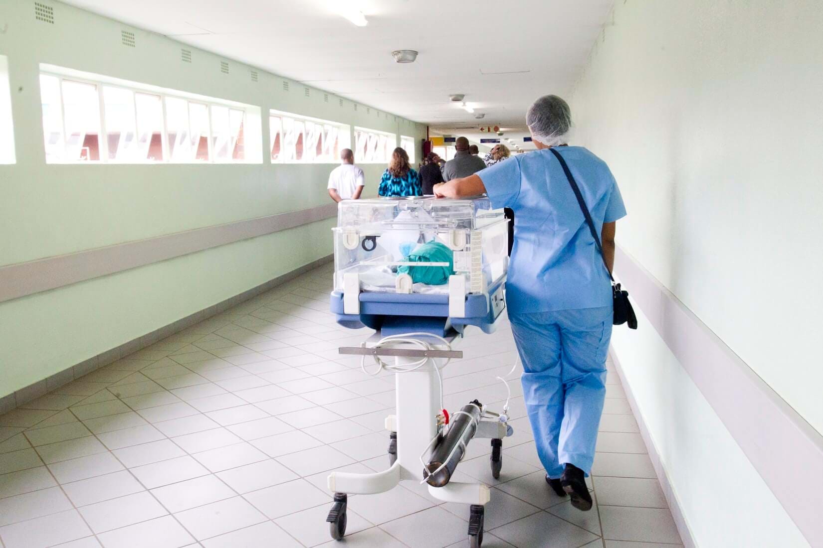 View of Person in Blue Medical Scrubs from Behind Rolling Incubator Down Hallway