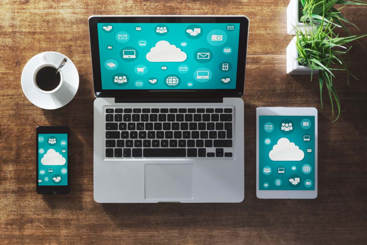 A mobile phone, laptop, and tablet all displaying a cloud surrounded by different icons over a greenish-blue background.