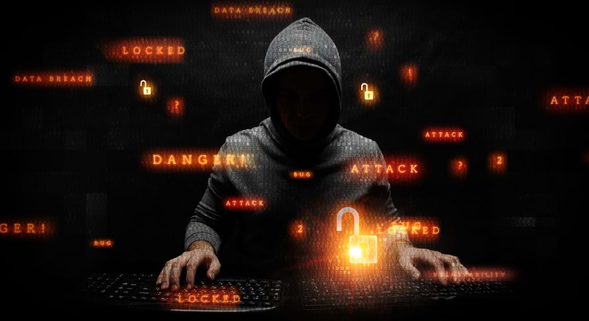 A hooded figure typing on two keyboards, with orangish-red text floating around him indicating he is committing cybercrimes