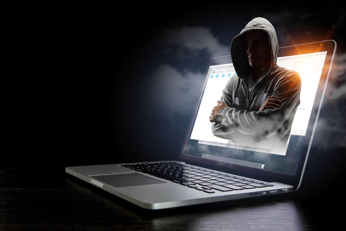 A laptop with a man with his arm crossed and their face obscured by a hoodie coming out of a laptop.