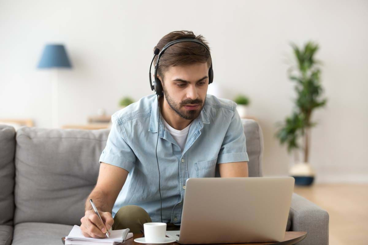 A man working from home using a laptop, headset, and writing on a notepad