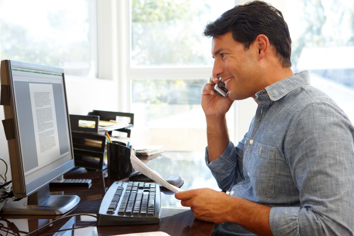 A man at his desk in his home office, talking on the phone while looking at the document open on his computer