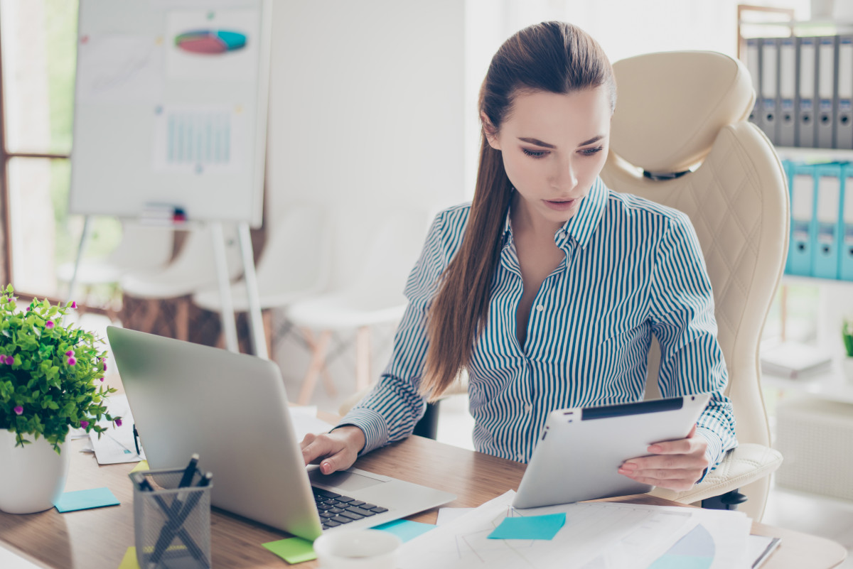 Businesswoman working on tablet and laptop computer seated at desk. 